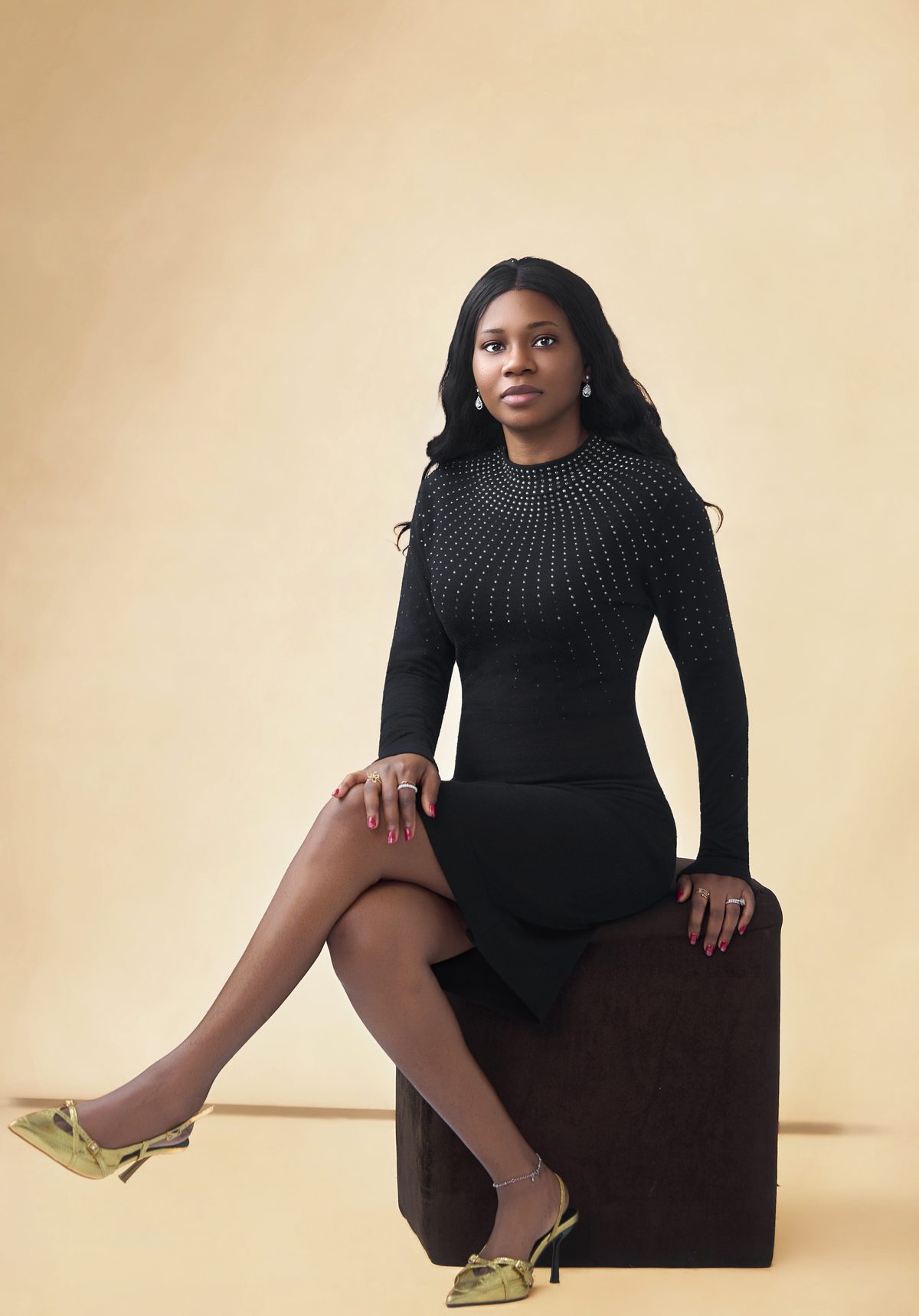 Seated editorial portrait in a black dress with gold heels.